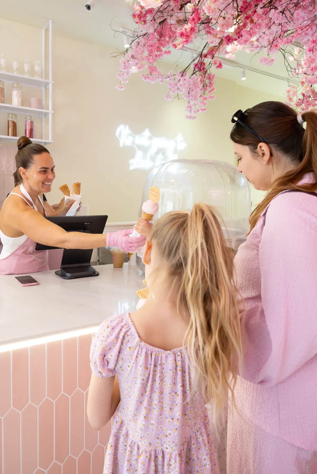 A member of the team handing a strawberry cone to a little girl at the counter, under the cherry blossom ceiling